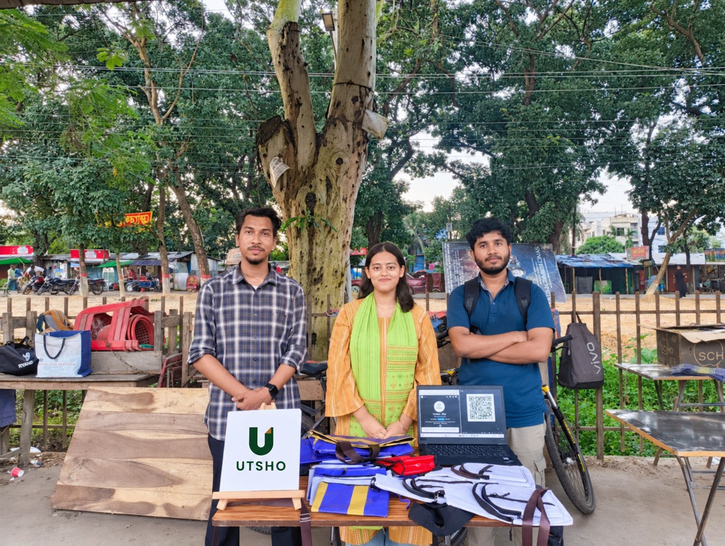 Three people behind a UTSHO stall displaying upcycled banner bags in Chattogram